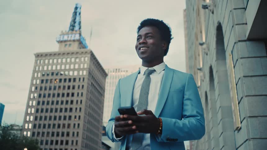 Man in business attire uses a smartphone in an urban area with buildings in the background
