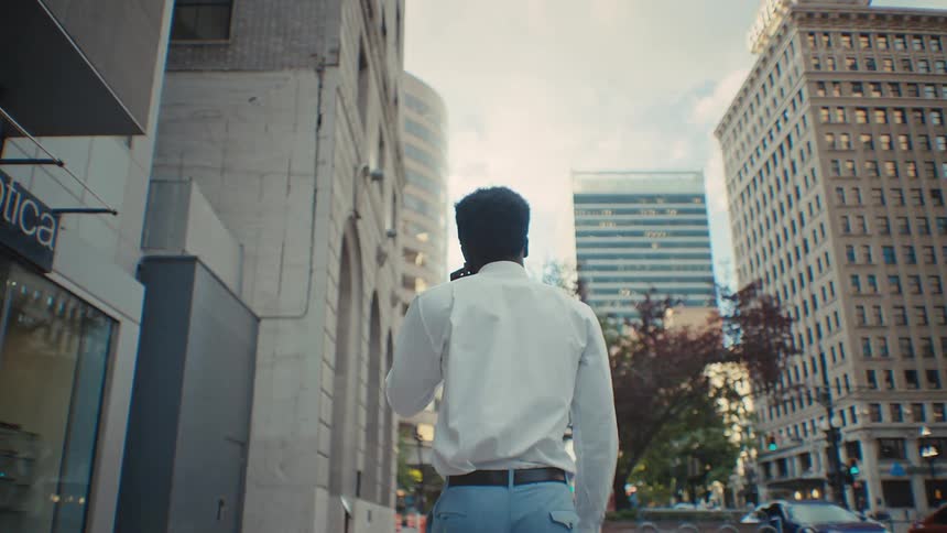 Black man wearing a white dress shirt and blue trousers walking on the sidewalk of a metropolitan area while talking on the phone outdoors during day time