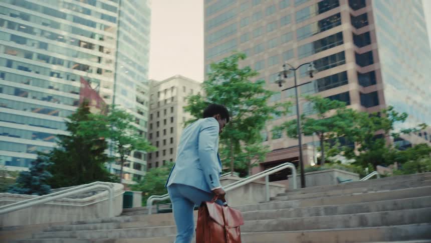 A young man walking up an outdoor staircase in the city at midday wearing a blue blazer suit and holding a brown suitcase answering a phone call as he climbs quickly