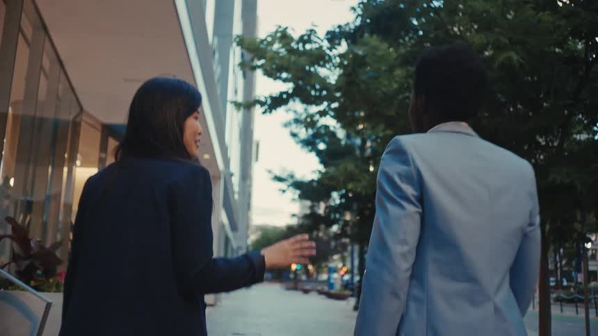 A young black man in a blue blazer suit walking beside a young asian woman in a black suit the two engaged in conversation as they move along a city sidewalk at midday