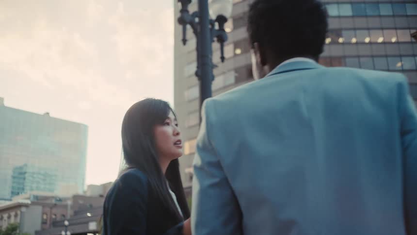 An asian businesswoman in a black blazer gestures while talking to a black male colleague in a light blue suit as they walk through a city downtown area during dusk surrounded by tall office buildings and street lights