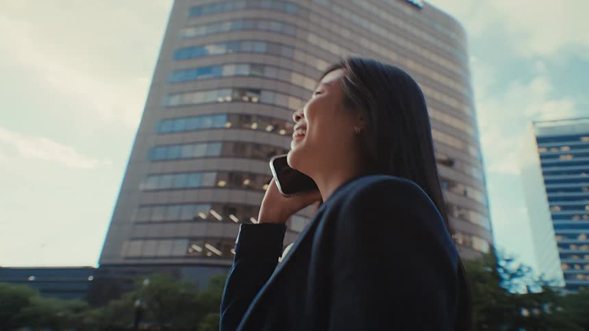 A smiling businesswoman in a dark blazer talks on her smartphone while walking past a large curved office building in a city