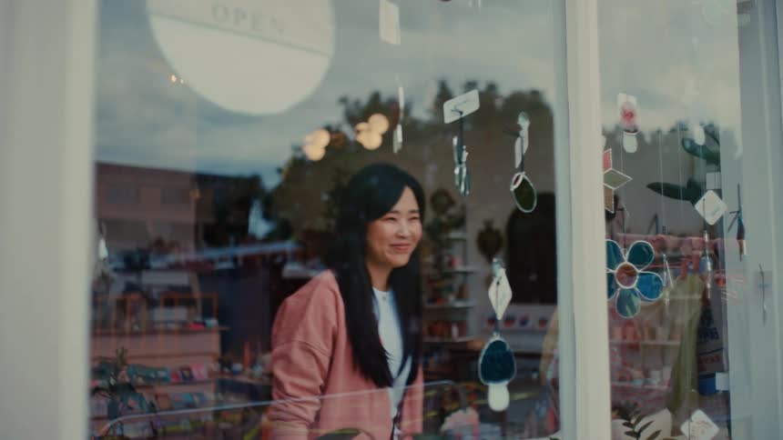 A smiling woman wearing a pink jacket looks through a glass storefront window decorated with colorful hanging flower ornaments