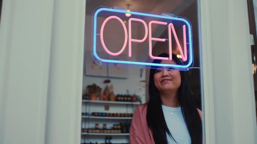 A retail shop owner smiles while turning on the shop lights as an open sign glows