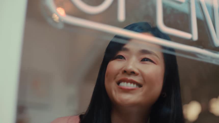 A young asian woman walking toward a store window at midday smiling as she looks at the open sign before stepping inside