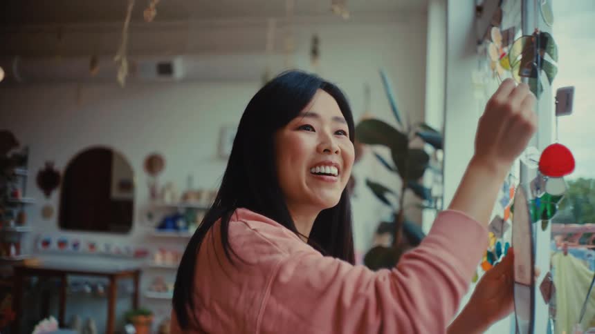 A young asian woman hanging an open sign on the stores glass window at midday smiling warmly as she finishes and gives a friendly wave to someone outside