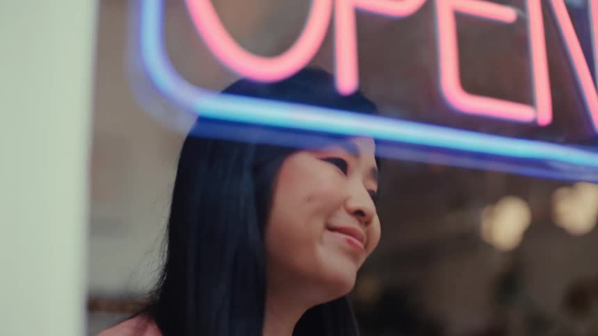 A smiling asian woman stands behind a glass storefront window as she turns on a pink and blue neon open sign then she looks out toward the street with a happy expression while blurred interior shop lights glow in the background