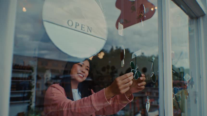 A smiling shop owner flips an open sign inside a retail storefront window