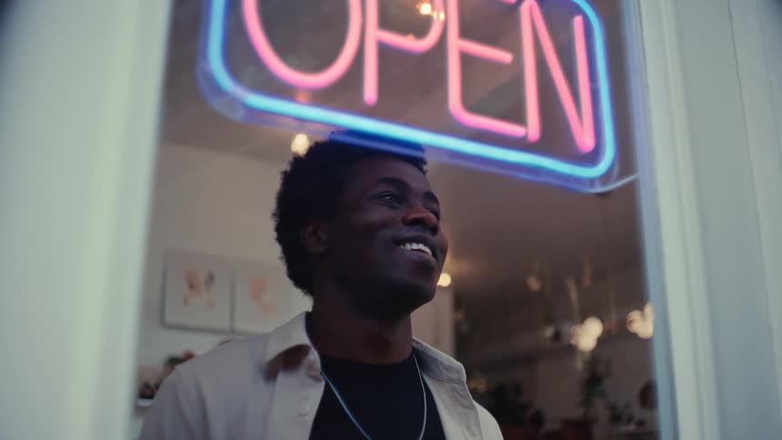 A young man stands behind a glass storefront and an open sign turns on with pink neon light