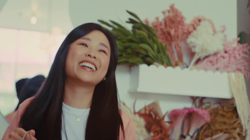 A customer completes a contactless payment with a credit card at a counter then a smiling woman in a pink cardigan hands them a brown paper shopping bag in a shop filled with floral arrangements and greenery