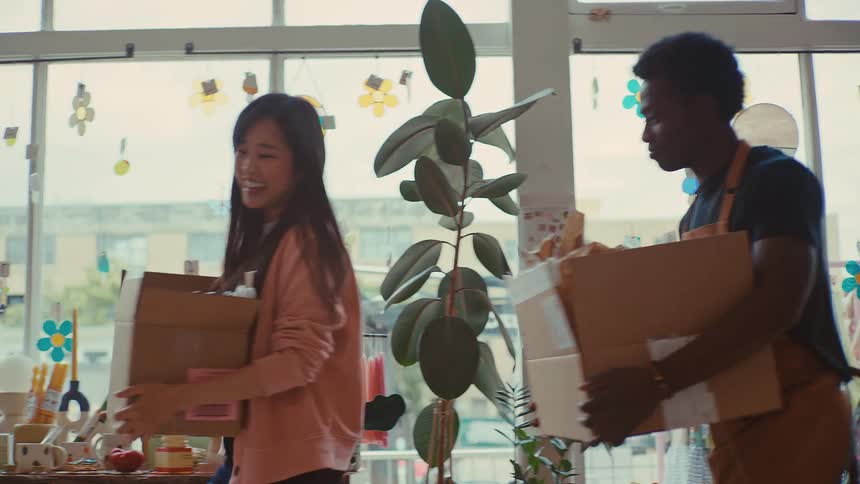 Two shop employees smile and chat while carrying boxes into a small retail store