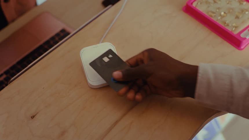 A person taps a black credit card on a white contactless payment terminal sitting on a light wooden counter next to a display of jewelry