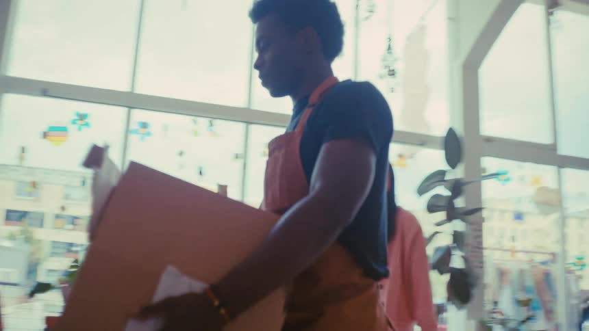 An asian woman points toward a shelf while a black man carries a cardboard box through a boutique filled with plants and clothes eventually bending down to place the box on a lower shelf near various ceramic items and home decor