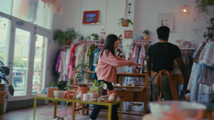 A male worker in an apron carries a cardboard box through a sunlit boutique filled with clothes and plants followed by a woman in a pink cardigan who helps him set the package down on a shelf in the back of the store