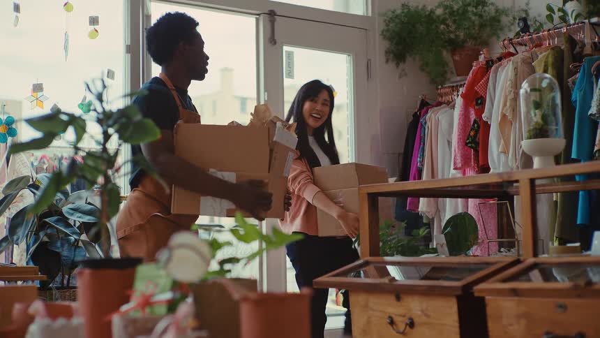 A man and a woman wearing aprons carry cardboard boxes into a bright clothing boutique while talking and laughing together