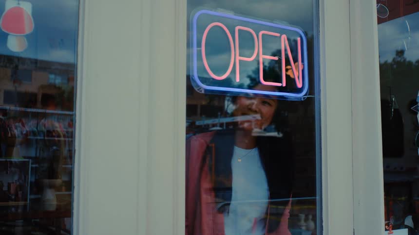 A retail shop owner turns on the lights inside her store as an open sign glows in the window