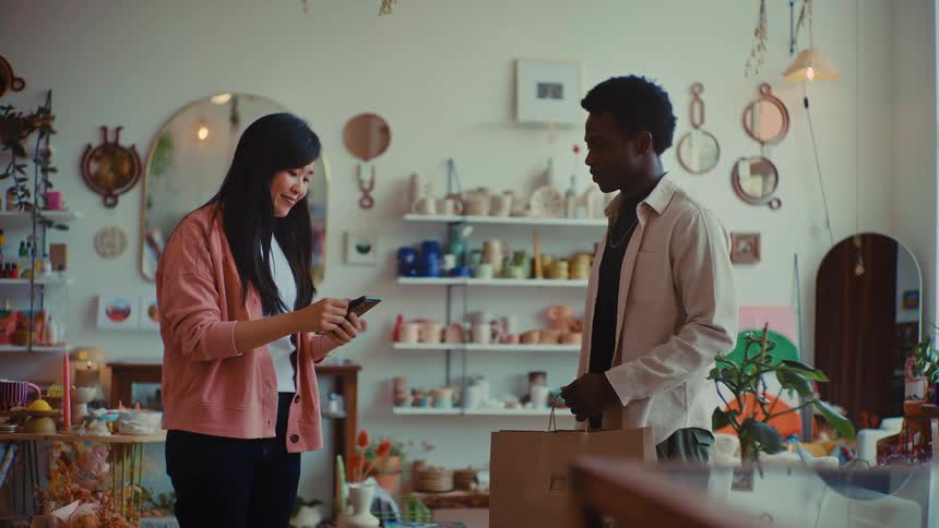 A smiling shopkeeper hands a paper shopping bag to a customer inside a warm artisan style store