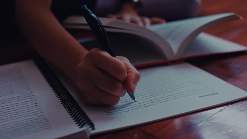 Woman writes in an open notebook while consulting a book on a wooden table