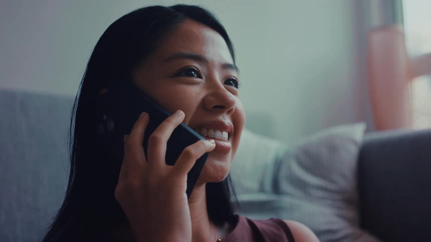 A smiling woman with long black hair speaks on a black smartphone while sitting indoors near a window with soft natural light