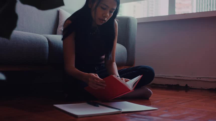 A young woman with long black hair wearing a brown sleeveless top and black pants sits on a wooden floor in a bright room and writes in a notebook while looking at an open red book