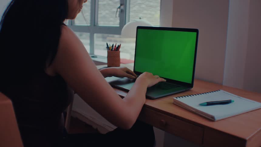 A woman sits at a wooden desk and types on a laptop and a notebook pen and container of pencils sit nearby as light shines through a window in the background