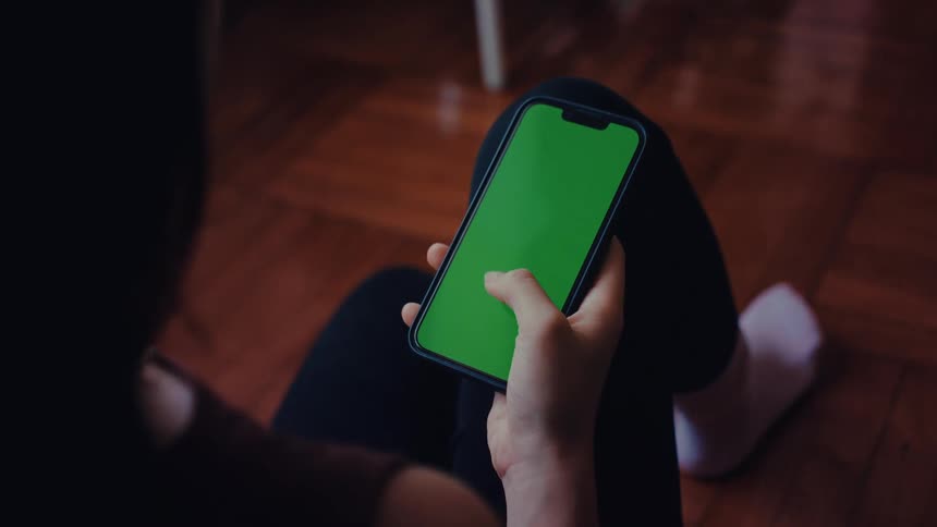A person holds a smartphone with a green screen while seated indoors