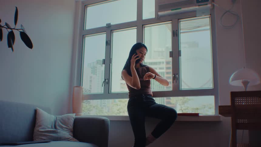 A woman wearing a brown sleeveless top and black pants sits on a wooden floor and uses her thumb to scroll through a smartphone with a bright green screen