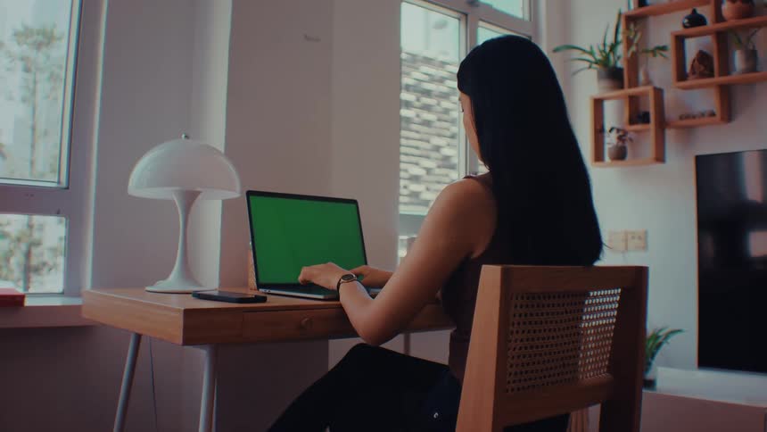 The woman is sitting at her desk and typing on her laptop