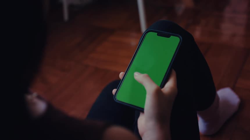 A woman wearing a brown sleeveless top and black pants sits on a wooden floor and uses her thumb to scroll through a smartphone with a bright green screen