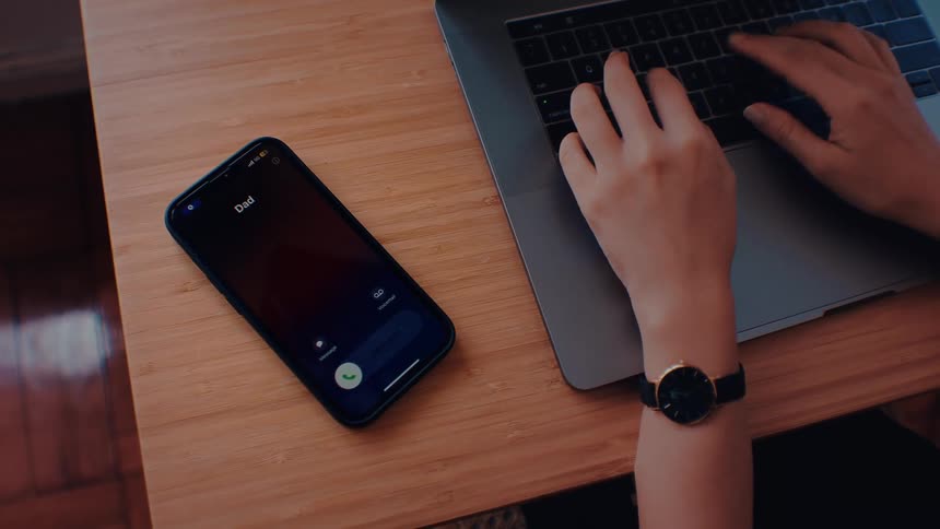 A person typing on a laptop receives a phone call from their dad and picks up the device