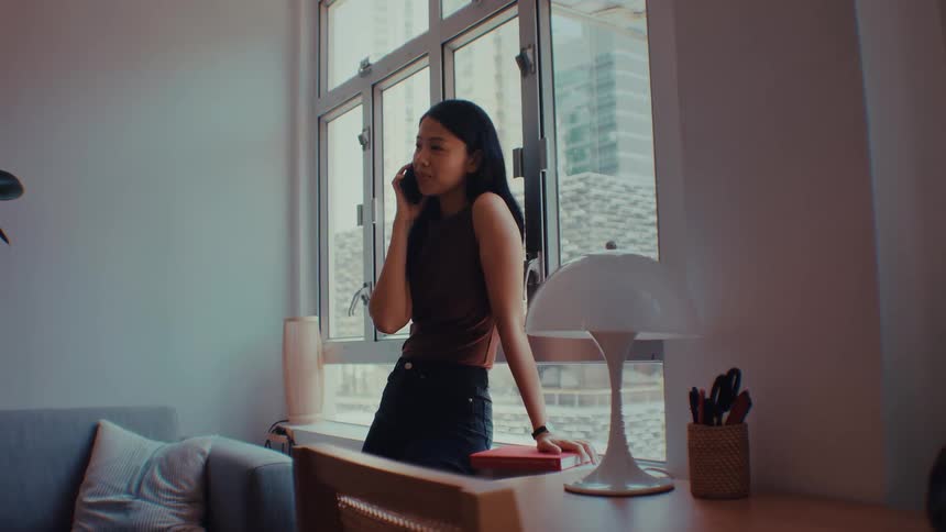 A young woman standing beside a window in her living room at midday answering a phone call while reaching for a redcovered book resting on the desk next to her