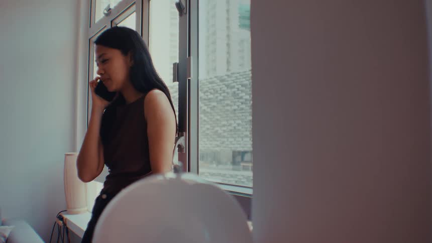 A smiling woman leans against an open window in an apartment while talking on a smartphone