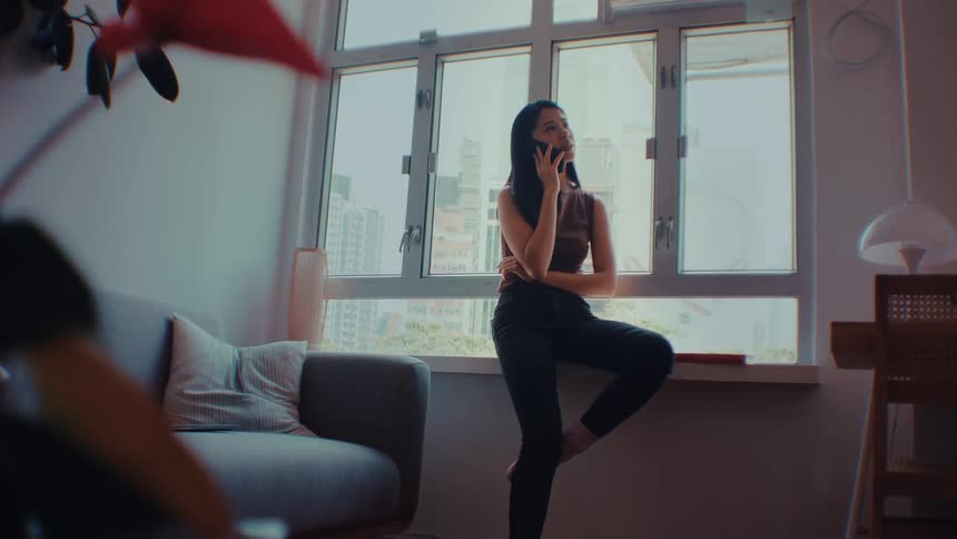 A woman leans against a windowsill in a sunlit apartment while talking on her smartphone and looking out at the city buildings