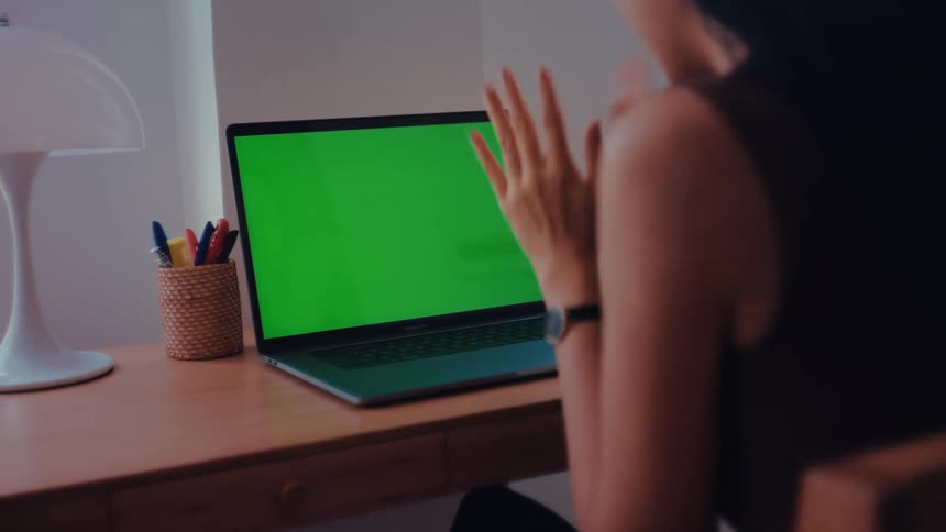 Teenage girl sitting at a table in front of a laptop computer with a green screen as she moves her hands and stops to think