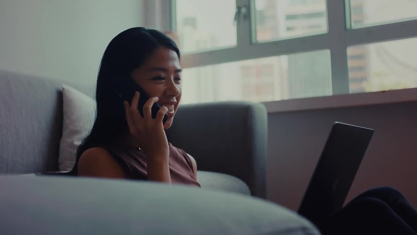 Teenage girl sitting on the living room floor in front of the couch typing on a laptop computer when she stops to answer her phone
