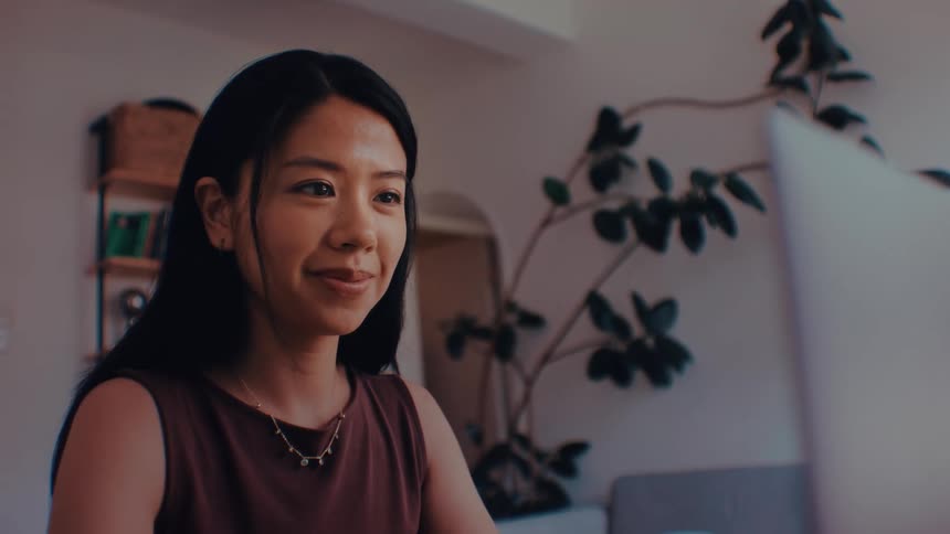 An asian teenage girl sits in a home office and looks at a laptop screen whilesmiling she has long dark hair and wears a brown sleeveless top