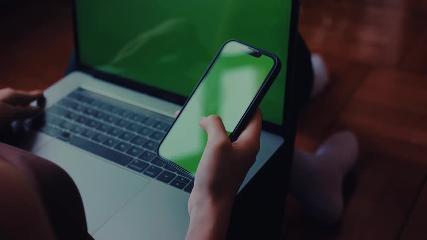 Teenage girl typing on a laptop computer as she stops to scroll on her phone