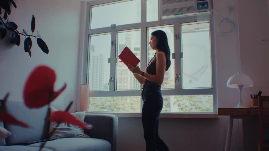 A young woman stands by a large window in a bright apartment thoughtfully reading a red book while city buildings are visible in the background