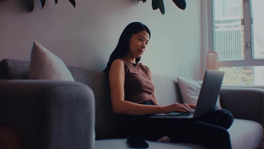 An asian woman sits on a grey couch in a living room and types on a laptop while looking at the screen