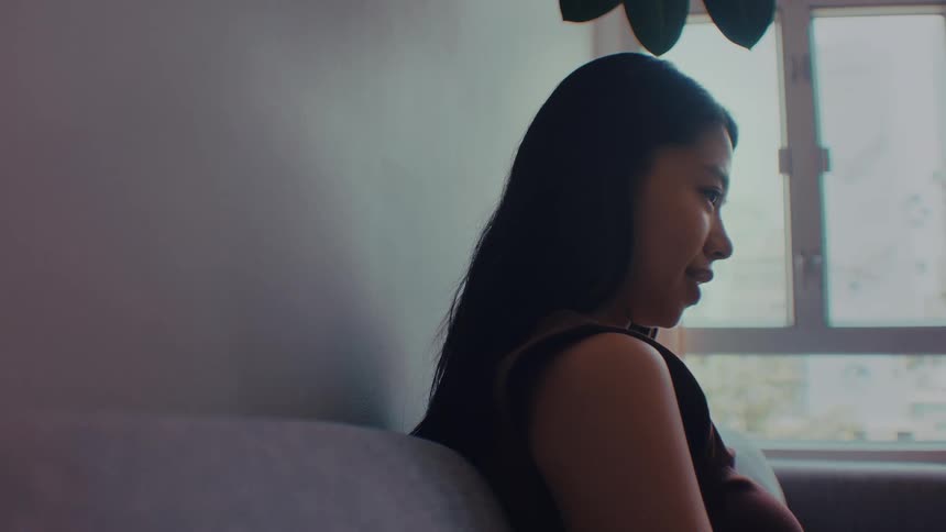 A young woman with long black hair wearing a brown sleeveless top sits on a couch in a bright room and smiles during a conversation