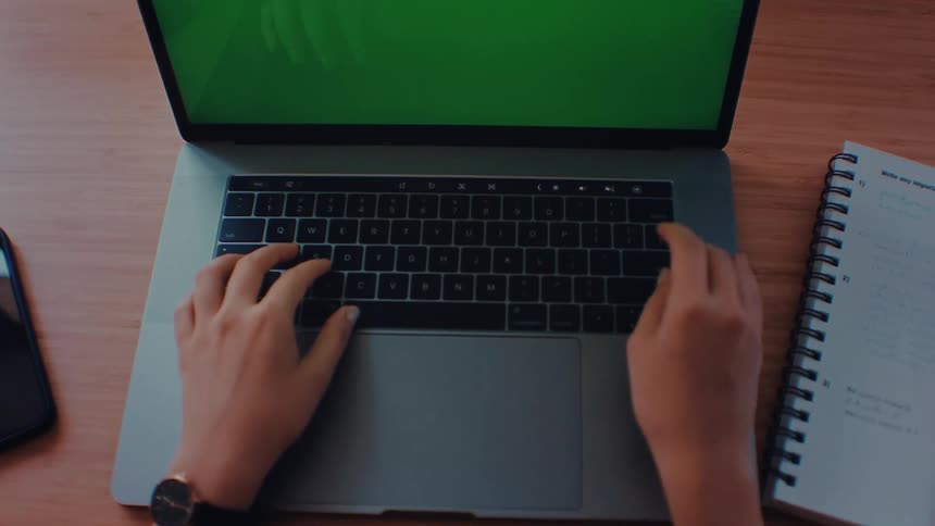 Person types on a laptop on a wooden desk with a notebook and smartphone nearby