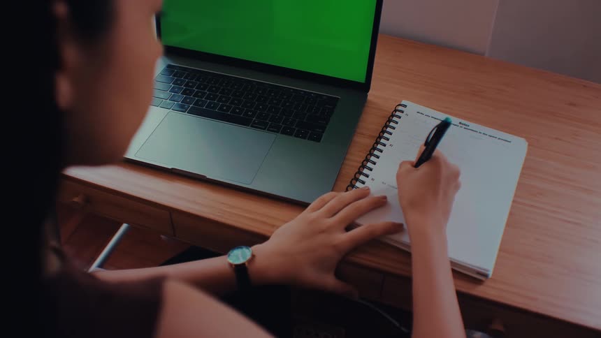 A person types on a laptop with a green screen before switching to write in a notebook