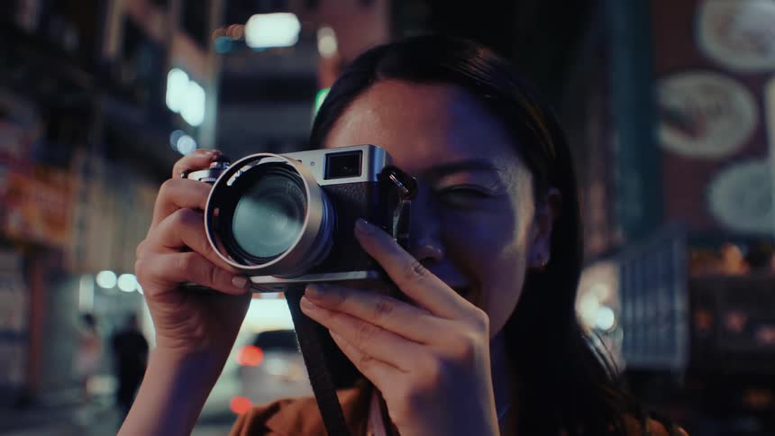 Woman stands on a city street at night smiling as she lifts her camera and takes a picture
