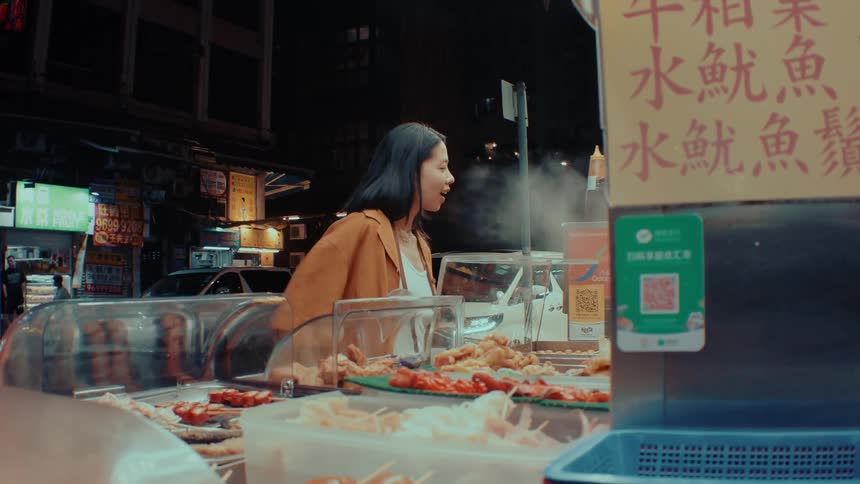 A young woman smiling as she makes a payment at a roadside food kiosk at midday holding out her method of payment while an elderly woman walks past on the sidewalk beside her
