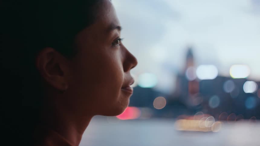 A woman looks out over a harbor at dusk as the city skyline begins to glow with lights in the background