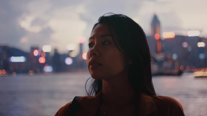 A young asian woman sitting outdoors with a calm waterway behind her and a city skyline rising in the distance