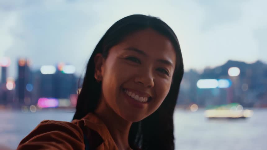 A woman with dark hair smiles warmly at the camera while taking a selfie outdoors at dusk behind her the blurred city lights of a skyline and harbor create a colorful bokeh background