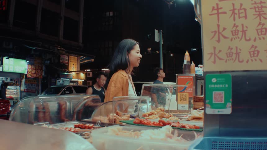 A young woman buying street food from a roadside vendor at night the scene lit by the vendors small lights as she waits for her order on the dimly lit street