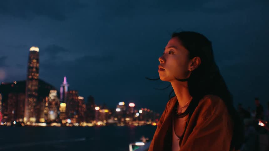 A young asian woman sitting outdoors with a calm waterway behind her and a city skyline rising in the distance at night time