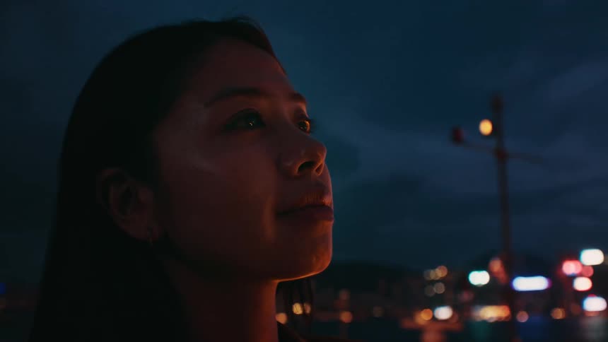 A woman stands in profile against a dark evening sky her face warmly illuminated by a nearby light sourcebehind her the soft glow of city lights creates a blurred multicolored bokeh effect over a body of water